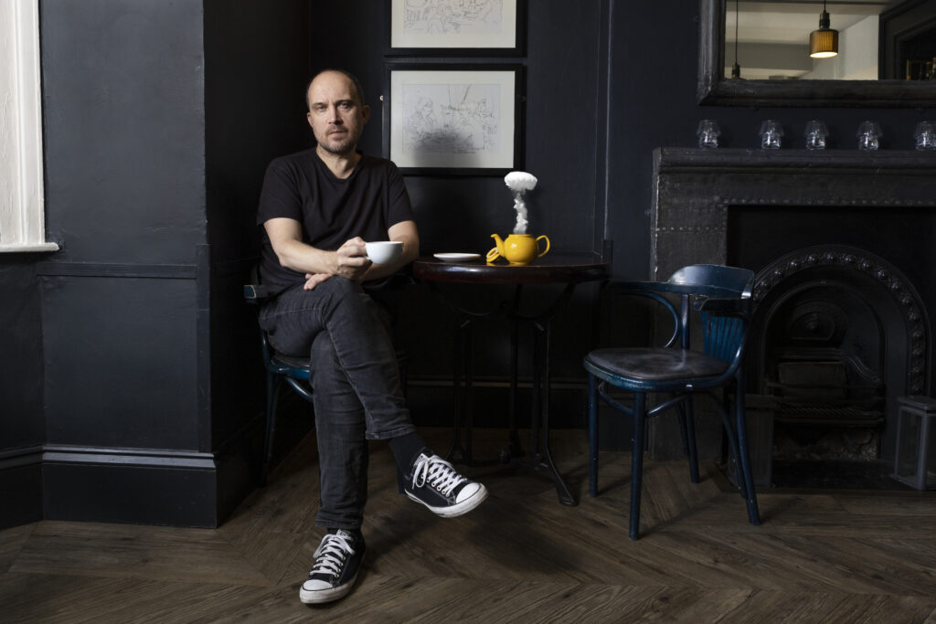 Image shows a man sitting cross legged on a chair, holding a mug, with a table, chair and fireplace to their left. On the table is a teapot with a large bloom of steam coming out.