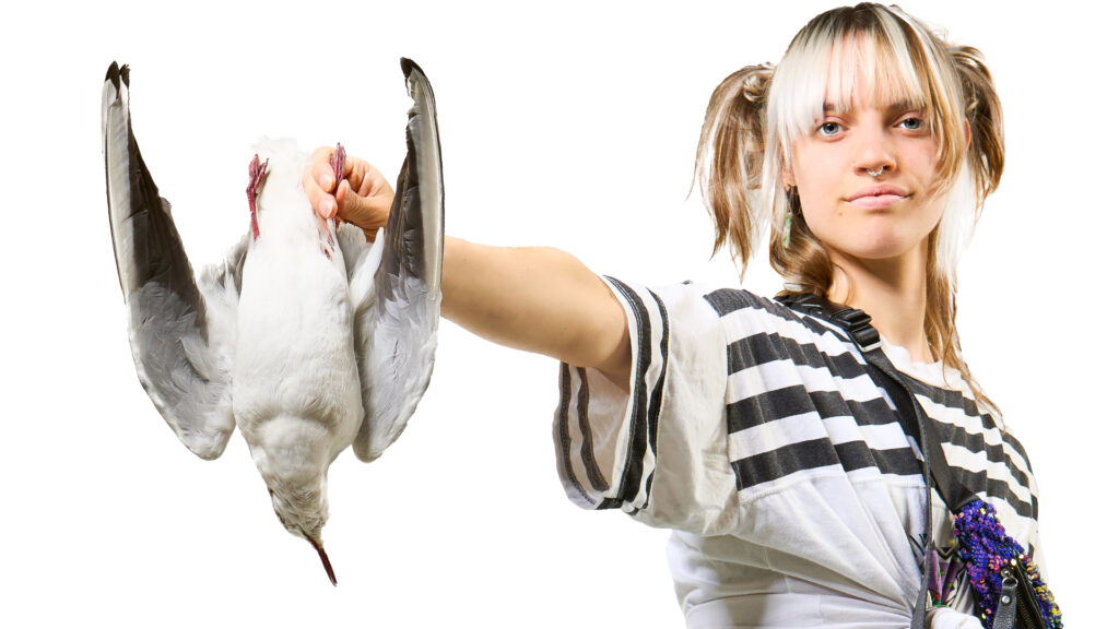 Image shows a woman with an outstretched hand, holding an upside down bird, by one foot.