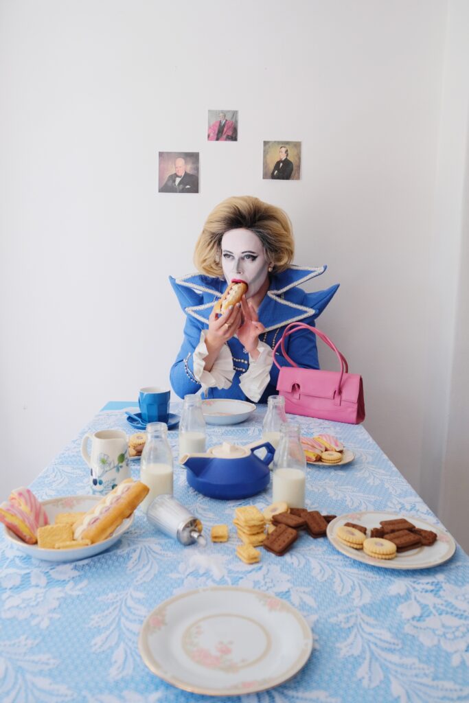 Image shows person dressed to look like Margret Thatcher, sitting at a table with a variety of cakes, biscuits, bottles of milk and a tea pot, with three small pictures behind them.