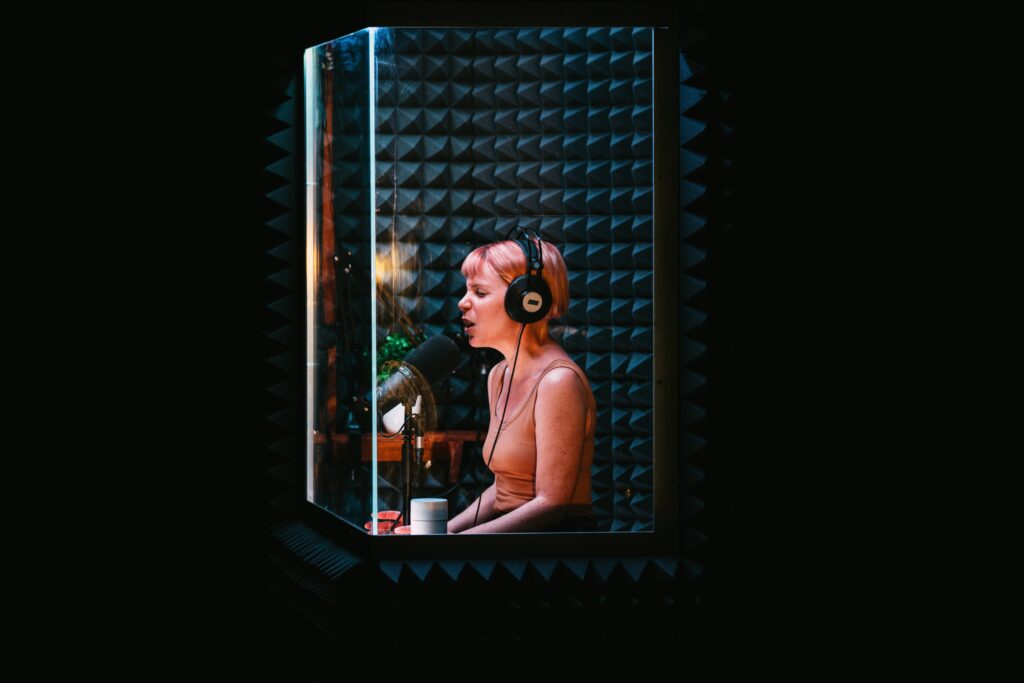 Image shows a woman in a small recording room, surrounded by sound dampening material, a microphone, a small table with a plant on top of it and a bottle to their left.