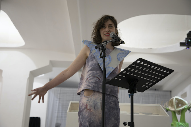 A photo of a dark haired woman standing in front of a microphone and speaking expressively 